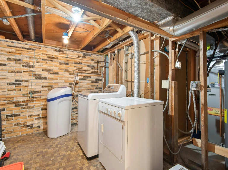 A basement laundry room with exposed wooden beams and pipes, a washing machine, dryer, water softener, and a brick-patterned wall. Some utility equipment and wiring are visible—ideal for those seeking expert plumbing in Toronto.