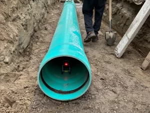 A close-up of a large green pipe lying in a trench during drain repair and plumbing in Toronto, with a device emitting a red light inside the pipe. A person stands nearby with a shovel, and a ladder rests against the trench wall.