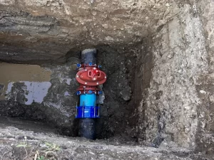 A large pipe with a red valve and blue connector, part of drain repair and plumbing in Toronto, is buried in a rectangular hole in the ground, surrounded by soil and a small puddle of muddy water.