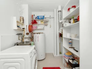 A small laundry room with white walls, a washing machine, water heater, shelves for cleaning supplies and towels, a utility sink perfect for drain repair and plumbing in Toronto, and a red rug. Pipes and ventilation are visible on the back wall.