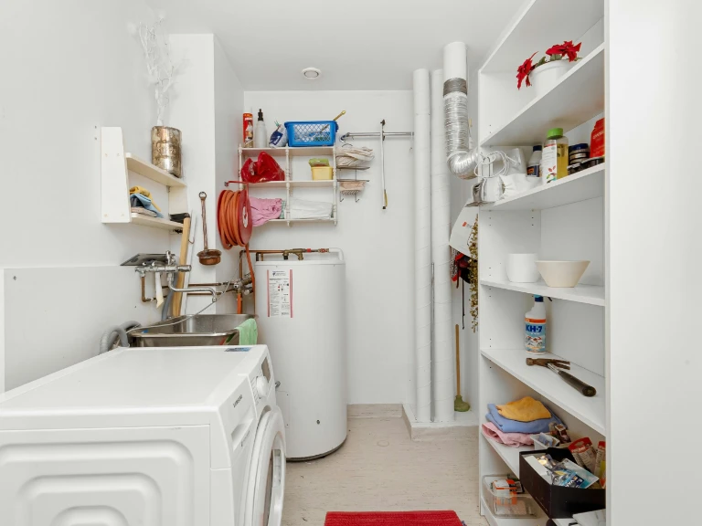 A small laundry room with white walls, a washing machine, water heater, shelves for cleaning supplies and towels, a utility sink perfect for drain repair and plumbing in Toronto, and a red rug. Pipes and ventilation are visible on the back wall.