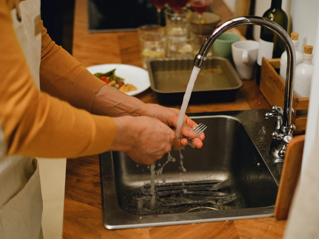 A person wearing a brown long-sleeve shirt is washing forks under running water at a kitchen sink. Dirty utensils are in the sink, and kitchen items are visible on the wooden countertop.