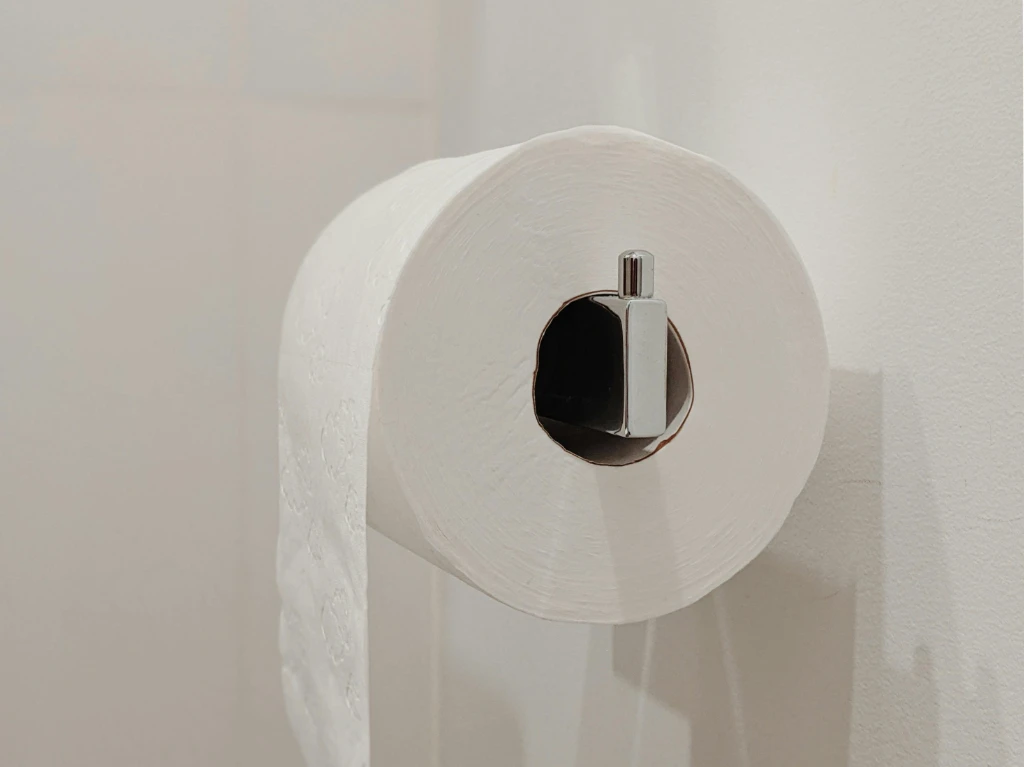 A large roll of toilet paper is mounted on a metallic holder attached to a plain white wall. One sheet hangs loosely from the roll.