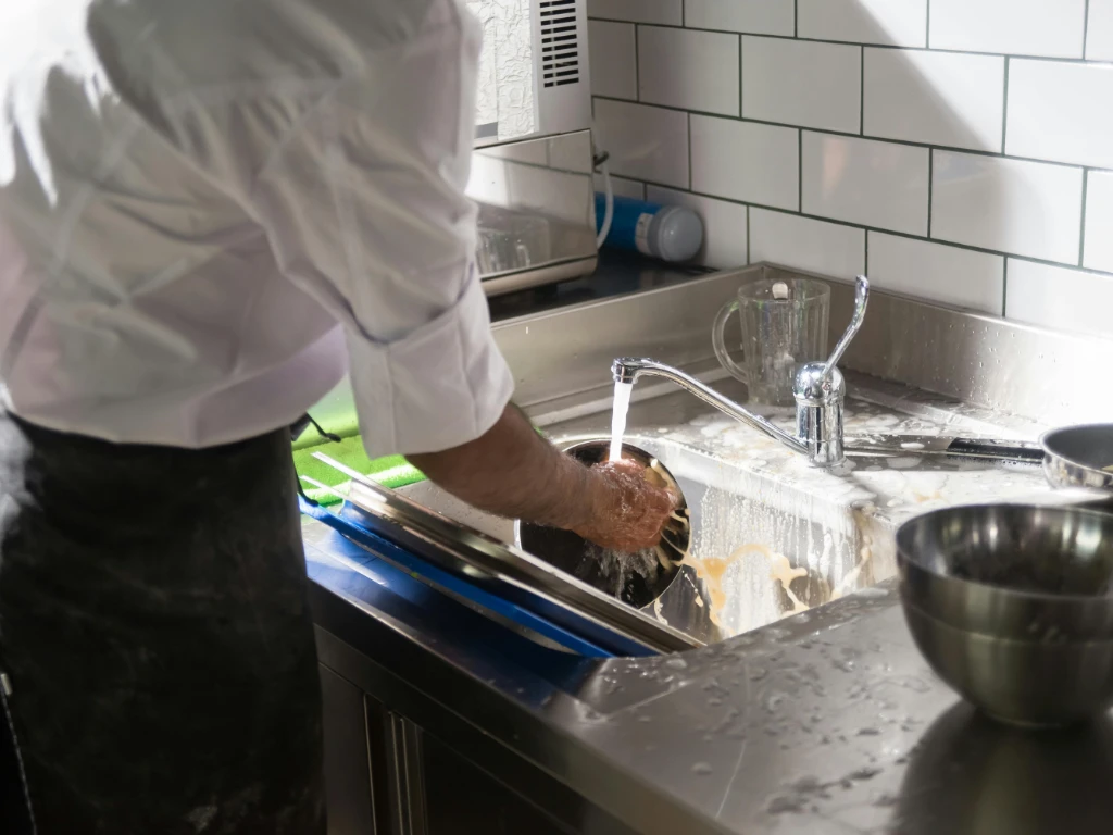 A person in a white shirt washes a pot under running water in a kitchen sink, with soapy water and metal bowls nearby on the counter.