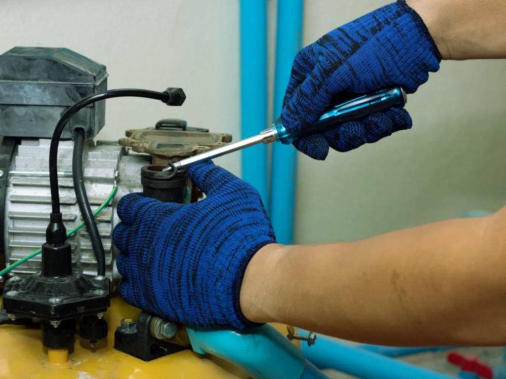 A person wearing blue work gloves uses a screwdriver to repair or adjust a machine with blue pipes and various wires attached.