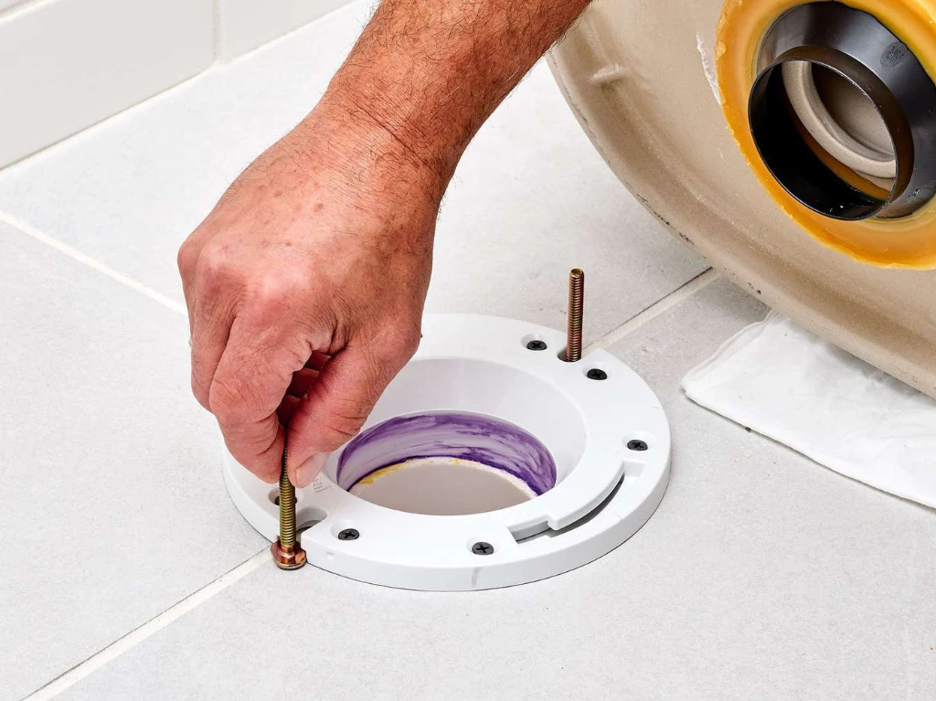 A person’s hand installs bolts into a white toilet flange mounted on a tiled bathroom floor, preparing to secure a toilet bowl in place.