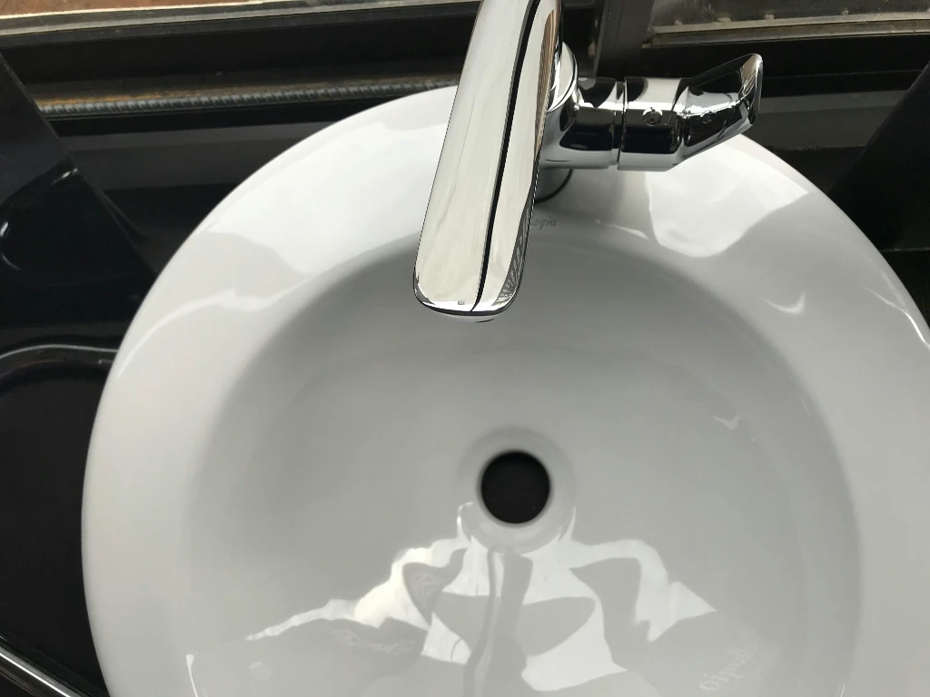 A close-up view of a white ceramic sink with a shiny chrome faucet, taken from above. The sink is clean and empty, with reflections of light visible on its smooth surface.