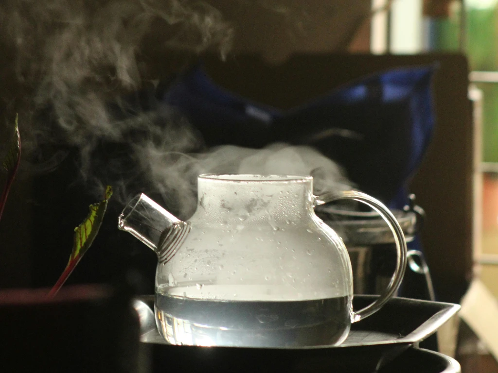 A clear glass teapot filled with hot water sits on a tray, with steam rising from its spout. The background is softly blurred, highlighting the warmth and coziness of the scene.