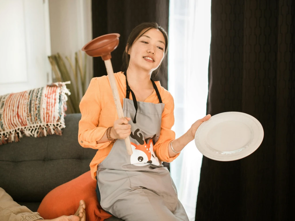 A woman wearing an apron sits on a couch holding a white plate in one hand and a plunger in the other, smiling. There are cushions and a blanket beside her, with curtains and a window in the background.