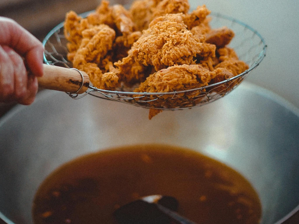 A hand holds a wire skimmer filled with crispy fried chicken pieces above a pot of hot oil, suggesting the chicken has just been cooked and is being lifted out of the fryer.