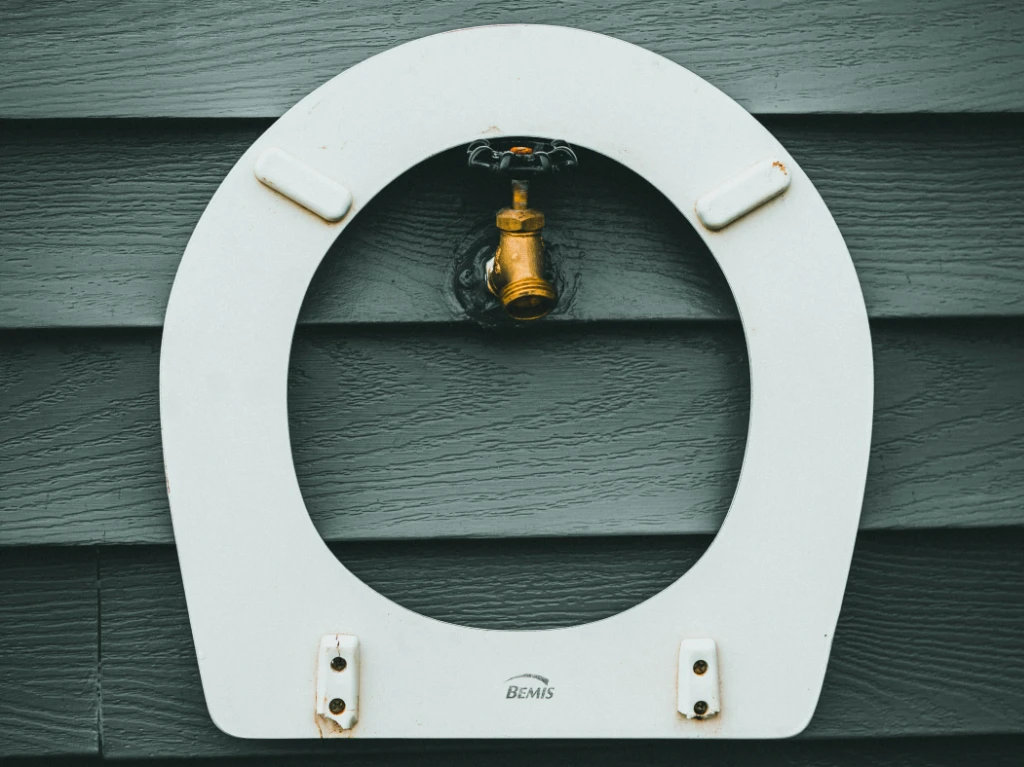 A white toilet seat mounted on a gray wall with a brass outdoor water spigot positioned in the center of the seat.