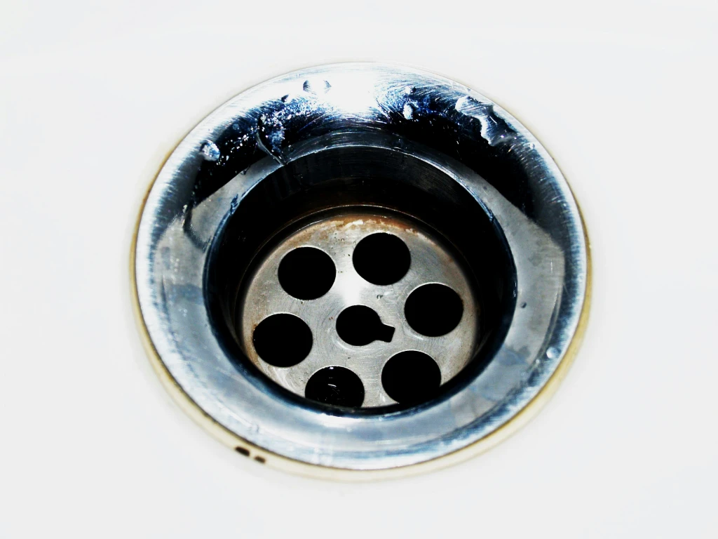 A close-up view of a shiny metal sink drain with six holes, set in a white sink. The metal surface reflects light and shows some water spots.