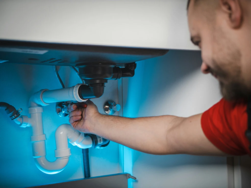 A person in a red shirt is repairing plumbing pipes under a sink, using a wrench to tighten a pipe connection. The area is illuminated with blue lighting.
