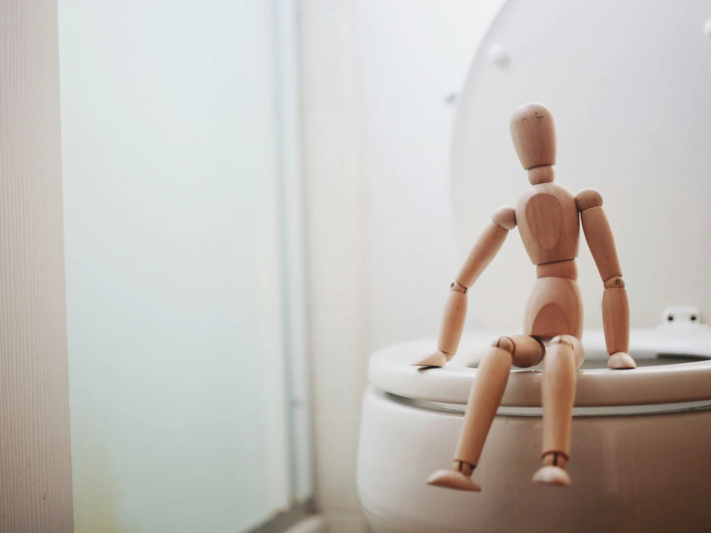 A wooden artist mannequin sits on a closed toilet lid in a bathroom, leaning slightly back with its legs dangling over the edge. The background is softly lit with a frosted glass window.