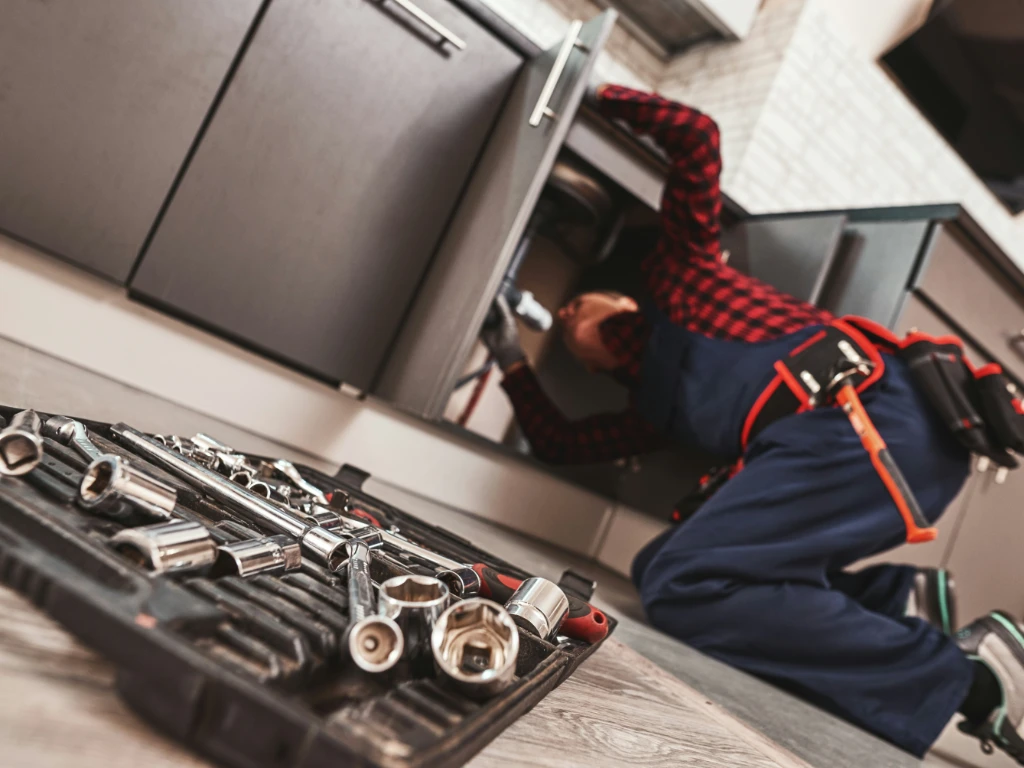 A plumber in a red plaid shirt and blue overalls kneels on the kitchen floor fixing pipes under a sink, with an open toolbox of metal wrenches and sockets in the foreground.