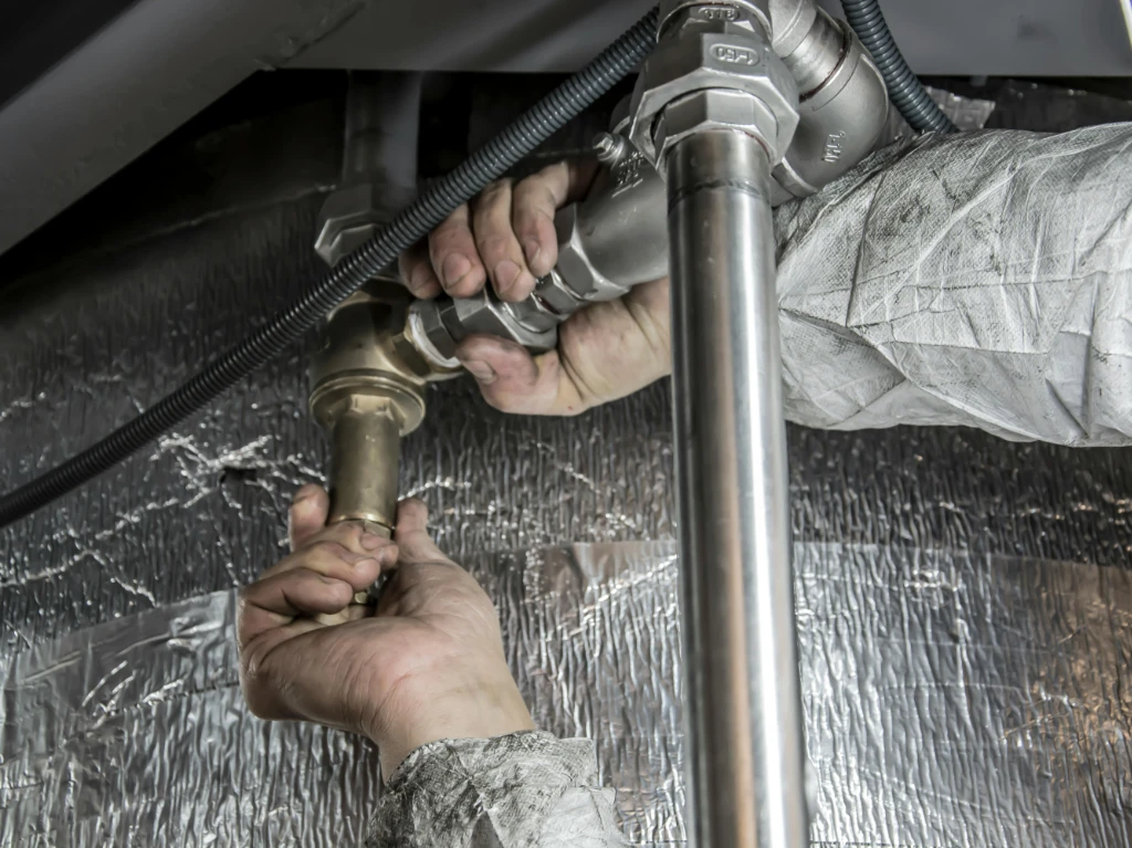 A person wearing protective clothing uses both hands to tighten a metal pipe fitting beneath a ceiling, with insulated and metal-wrapped surfaces in the background.