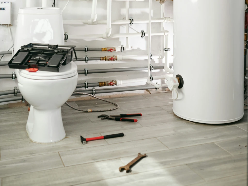 A bathroom with plumbing tools scattered on the floor, a toilet with a tool box on top, and a white water heater nearby; pipes and valves are visible along the wall.