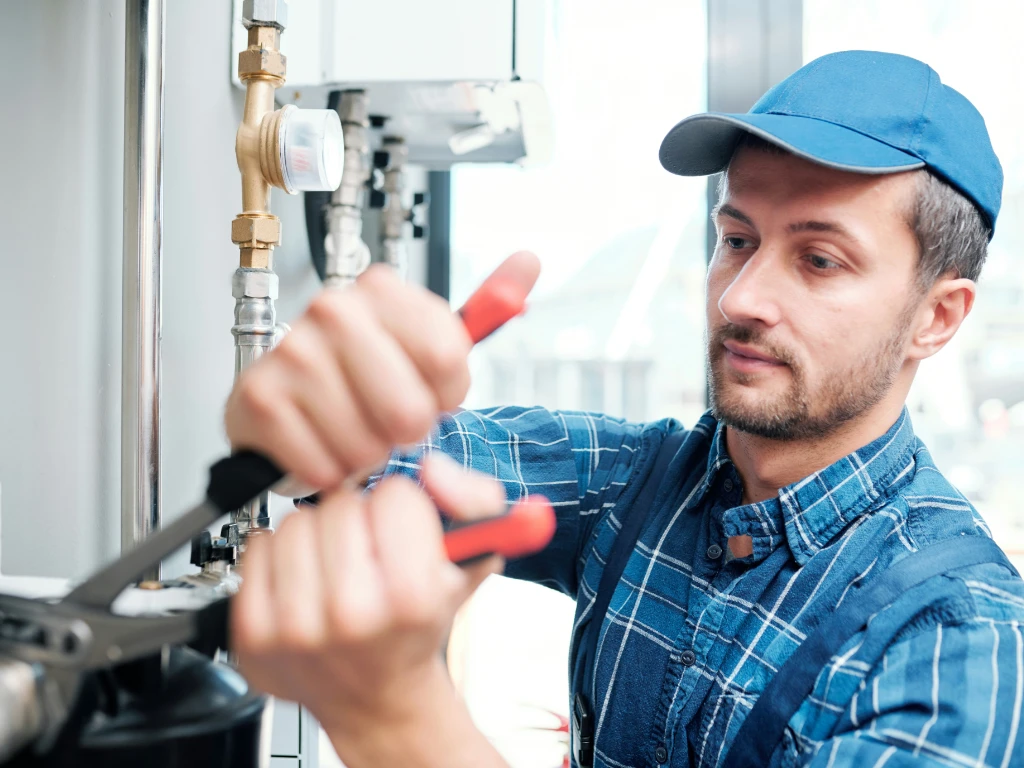 A man wearing a blue cap and plaid shirt uses pliers and a screwdriver to work on metal pipes and valves, appearing focused while performing plumbing or maintenance work indoors.
